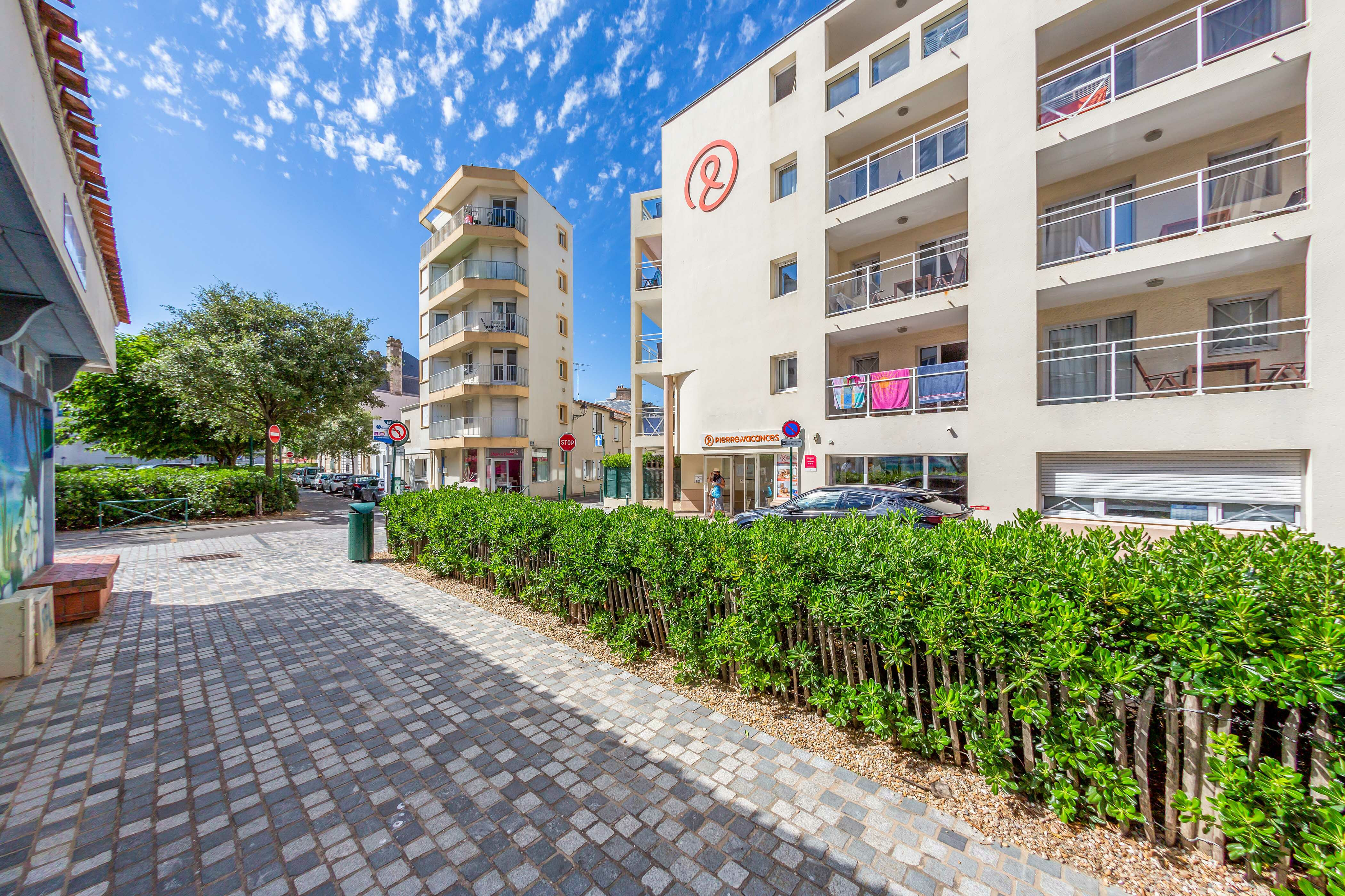 Apartment buildings with balconies surrounded by greenery and a cobblestone path under a bright blue, partly cloudy sky at La Baie des Sables
