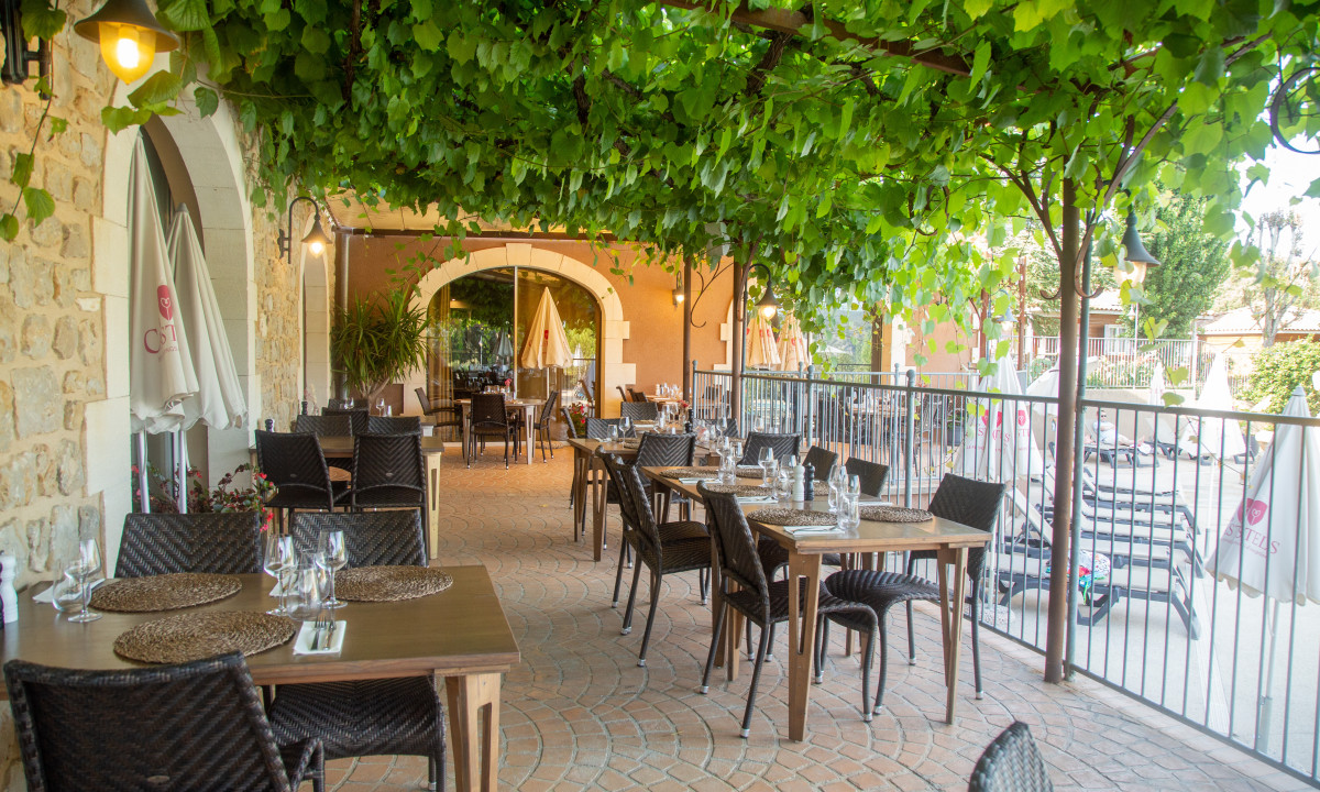 Tables set with placemats and glasses arranged under a pergola with vine leaves, adjacent to a stone building at Domaine de Sévenier