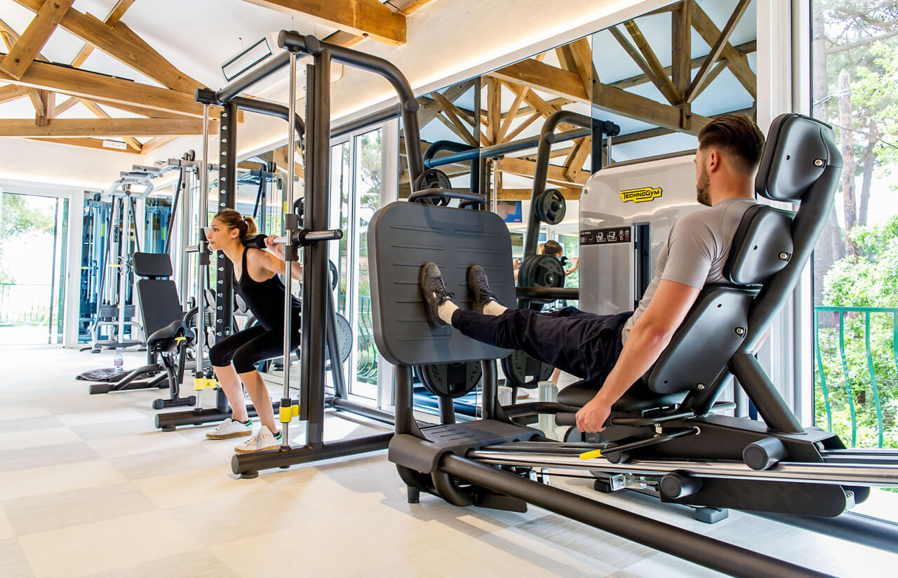 At Bois de Valmarie, individuals are working out on gym equipment within a brightly lit, contemporary fitness center, encircled by expansive windows that offer views of the forested landscape.