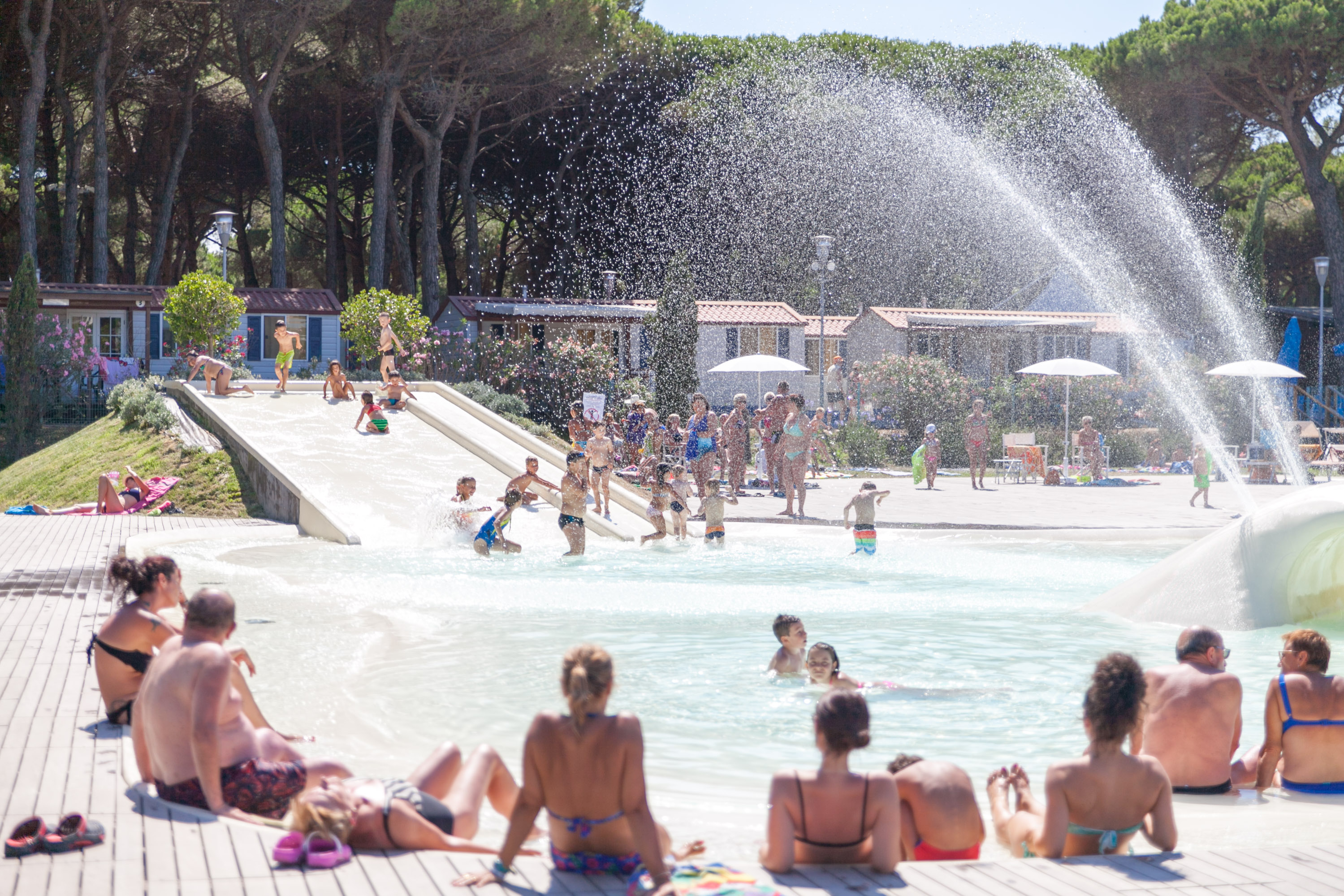 People lounging and kids playing in a water park near slides and sprinklers, surrounded by trees and cabins at Pineta Sul Mare Camping Village
