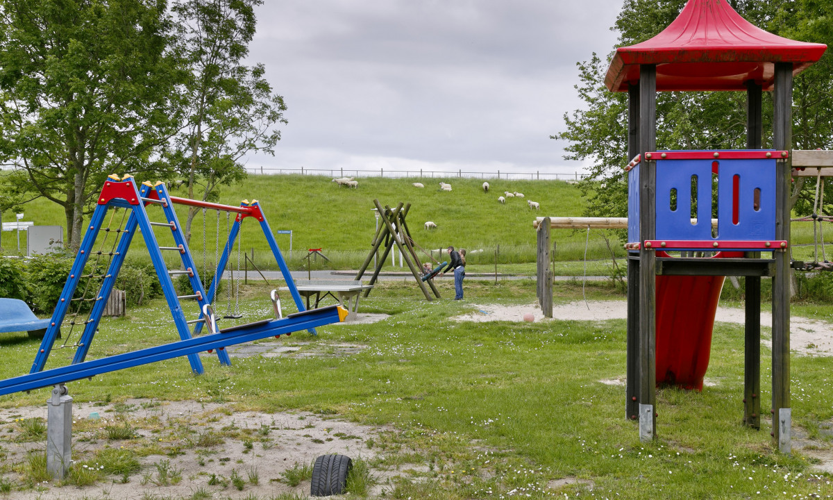 At KNAUS Campingpark Eckwarderhörne, a playground featuring slides and swings is filled with a child and an adult, encircled by grass, trees, and sheep in the distance.