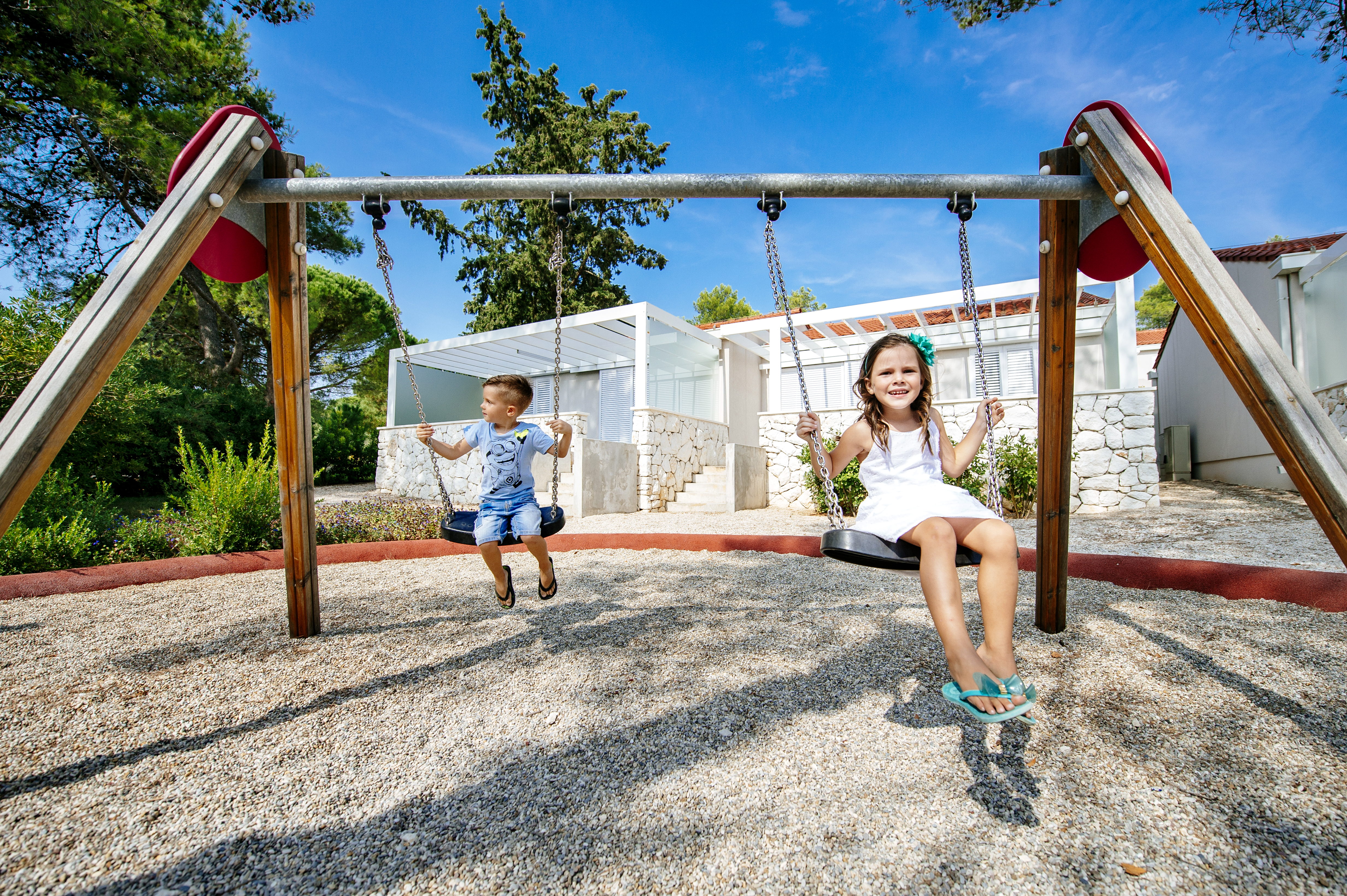 At Zaton Holiday Resort, kids swing happily on a playset, encircled by gravel, vibrant greenery, and contemporary structures.