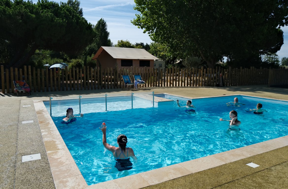 People swimming in a clear blue pool surrounded by trees and wooden fencing, with tents and lounge chairs nearby at Seasonova Ile de Ré