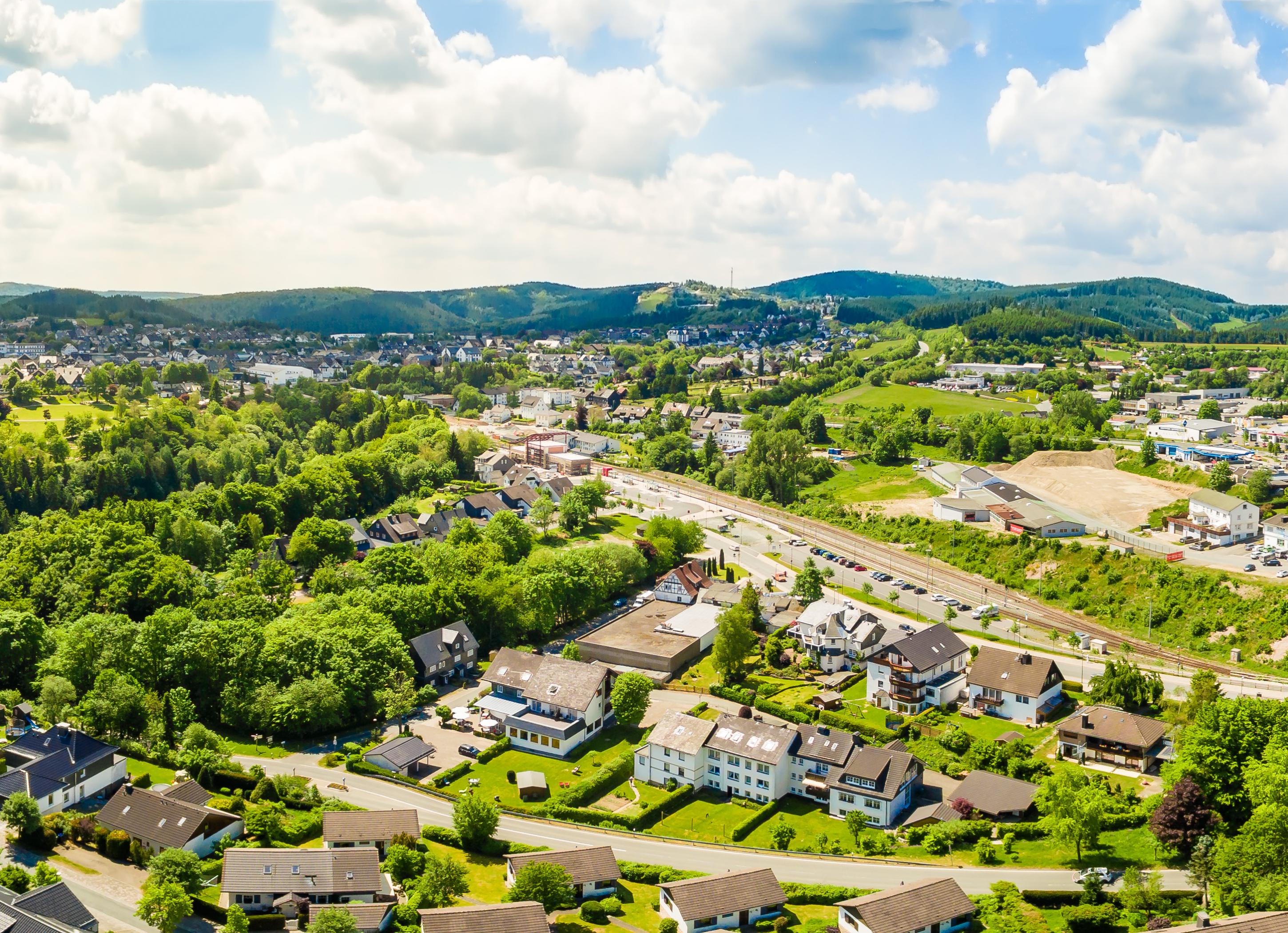 Aerial view: Small town surrounded by lush green hills and a railway, under partly cloudy skies at Bergresort Winterberg