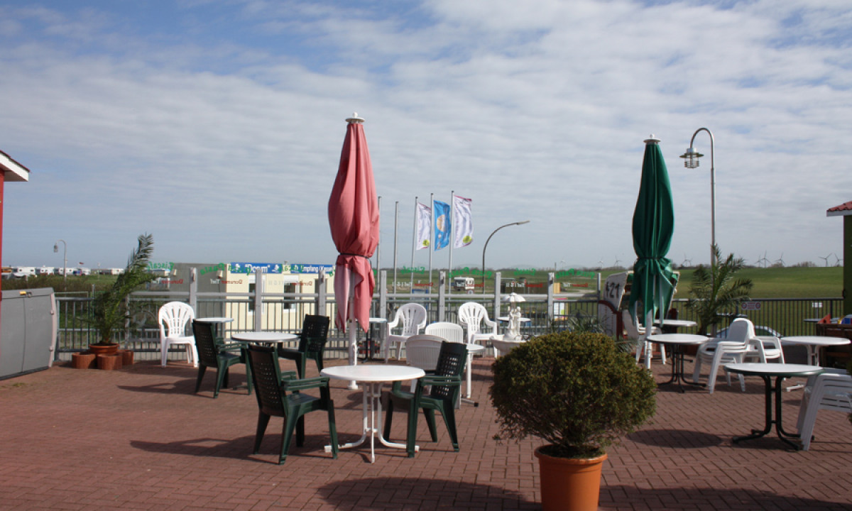 At KNAUS Campingpark Dorum, there are seats and tables surrounding folded umbrellas on a brick terrace, with grassy fields and flags visible in the background.