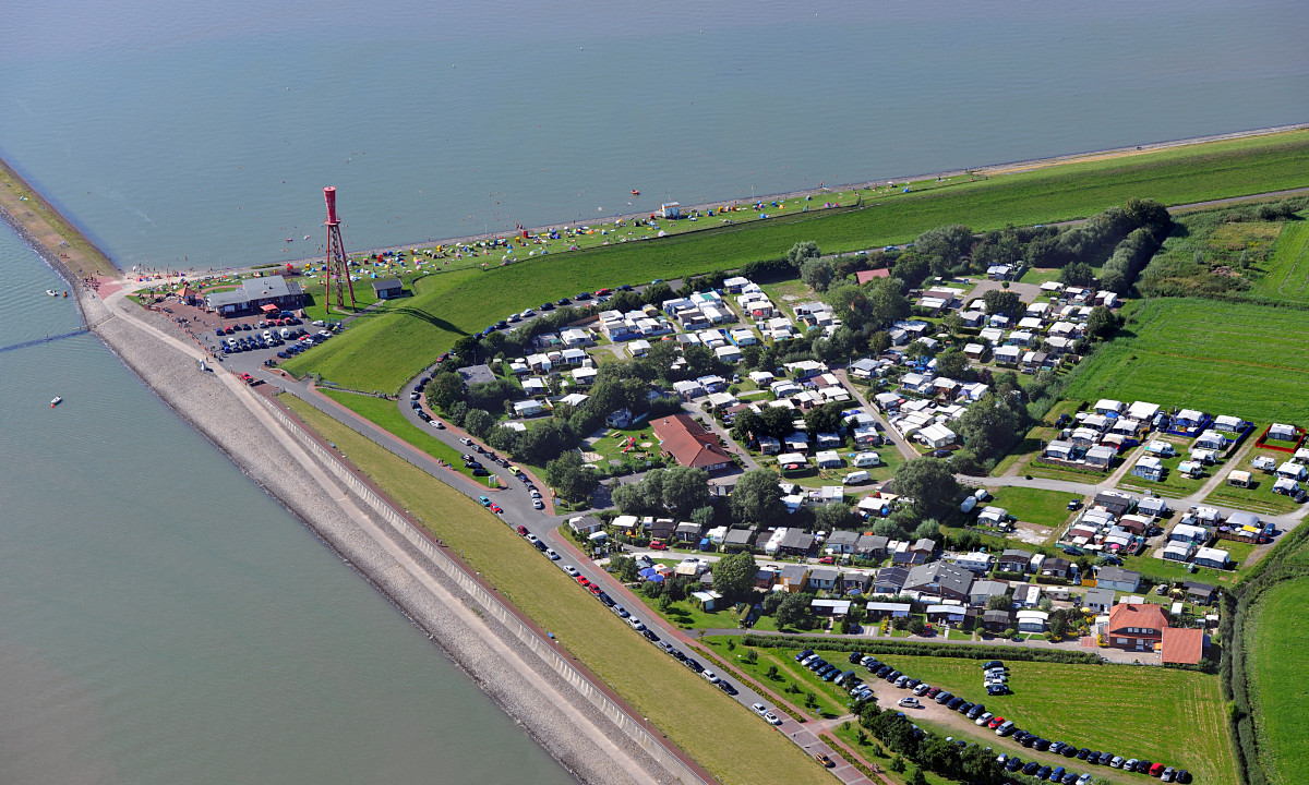 At KNAUS Campingpark Eckwarderhörne, an aerial perspective reveals a camping area close to the shoreline, showcasing a variety of tents and recreational vehicles, alongside a lighthouse.