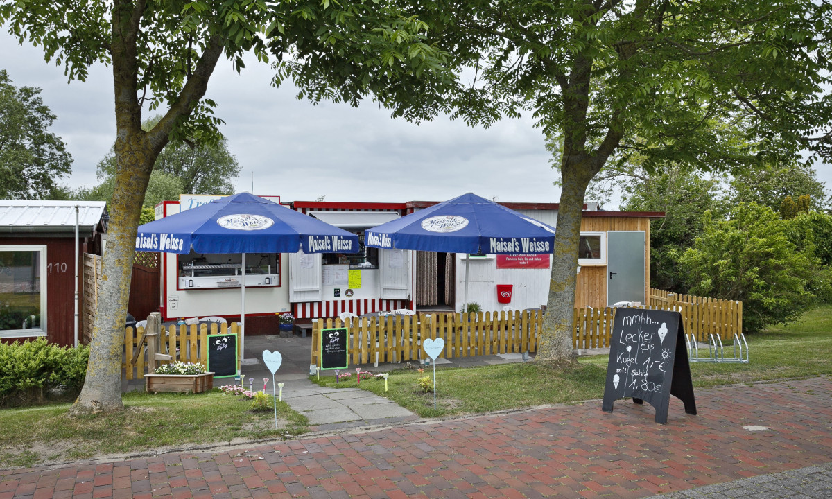 At KNAUS Campingpark Eckwarderhörne, a small food stall offers ice cream, featuring blue canopies, a wooden enclosure, lush greenery, and a brick walkway.
