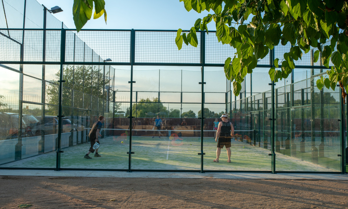 Two people playing paddle tennis in an outdoor court enclosed by mesh fencing, with trees and parked cars in view at Resort Els Pins