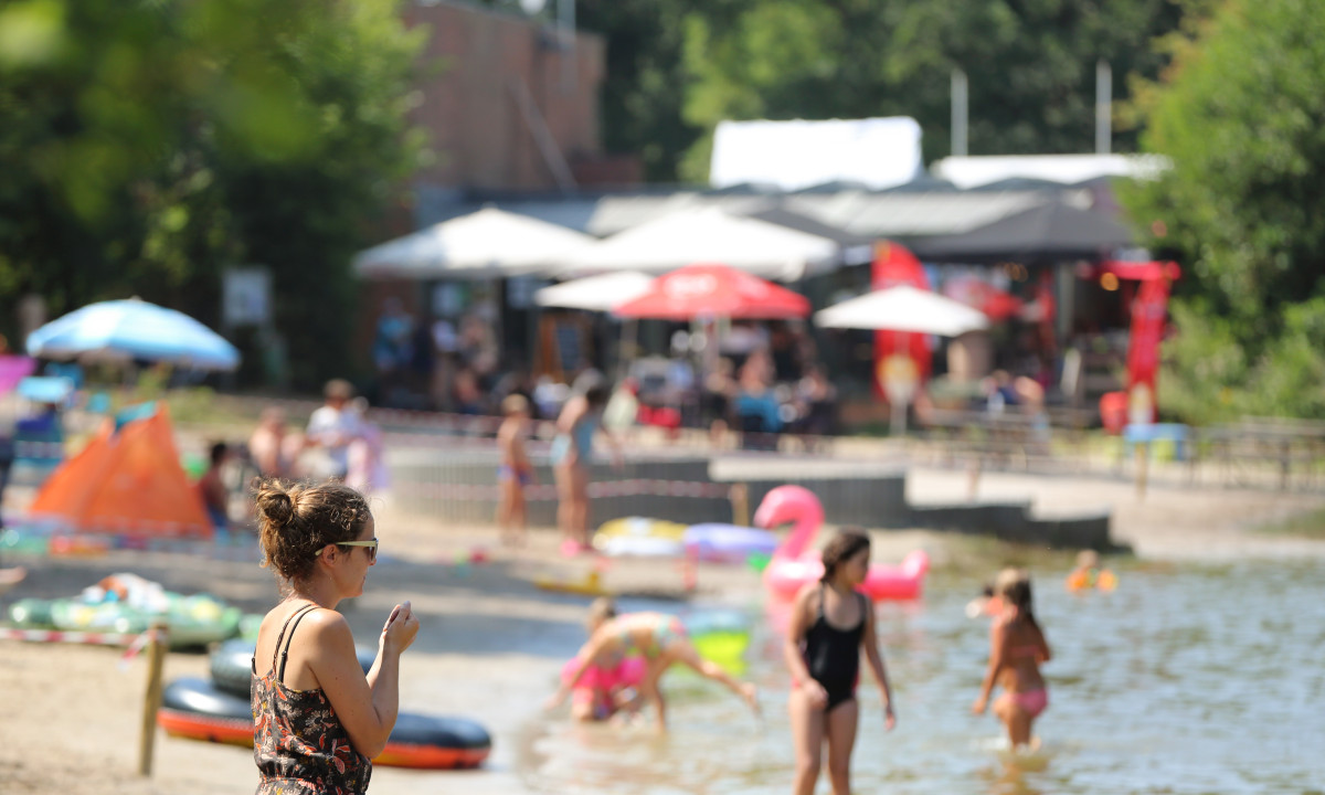 At De Baalse Hei, an individual stands on the shore, watching kids frolicking in the water with floatation devices, close to a lively outdoor café.