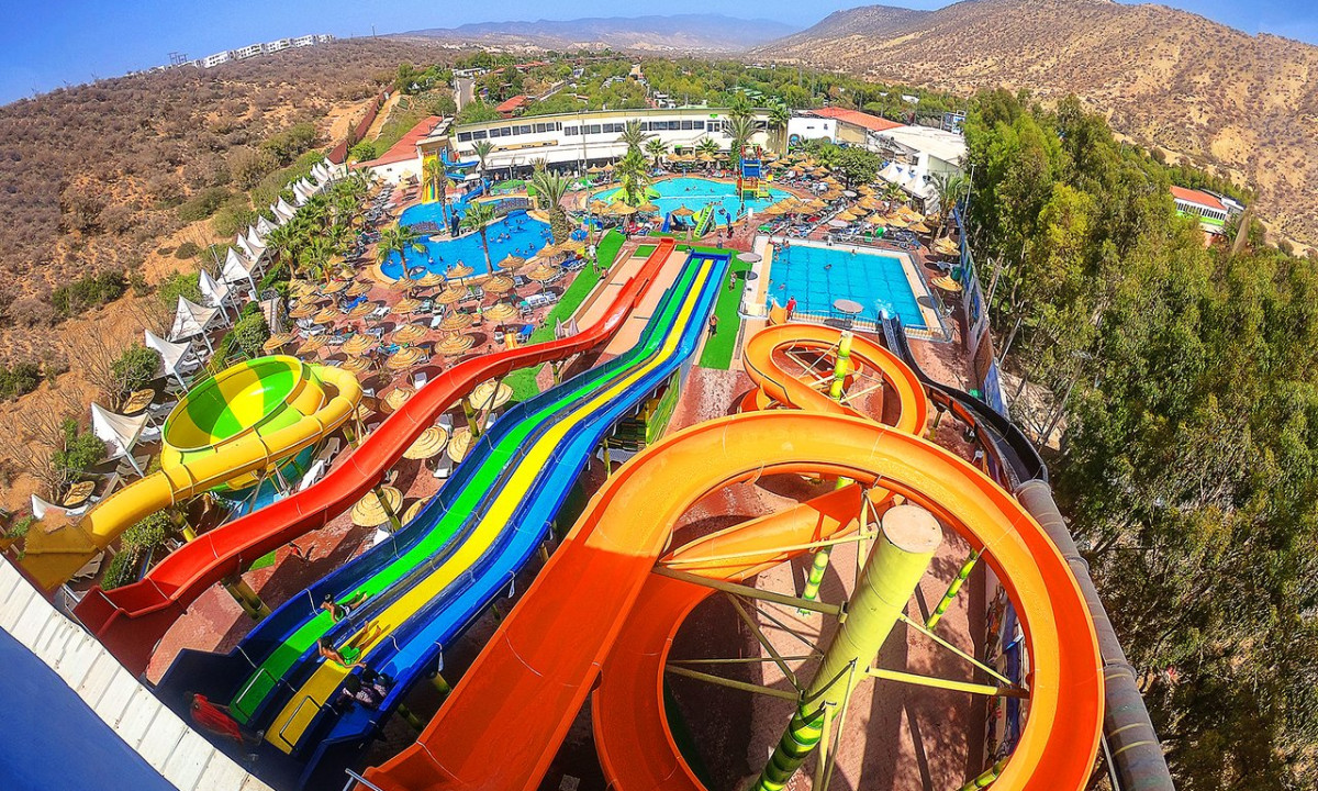 Water slides winding through an outdoor water park, surrounded by pools, umbrellas, and lounge chairs; hills in the background at Atlantica Parc