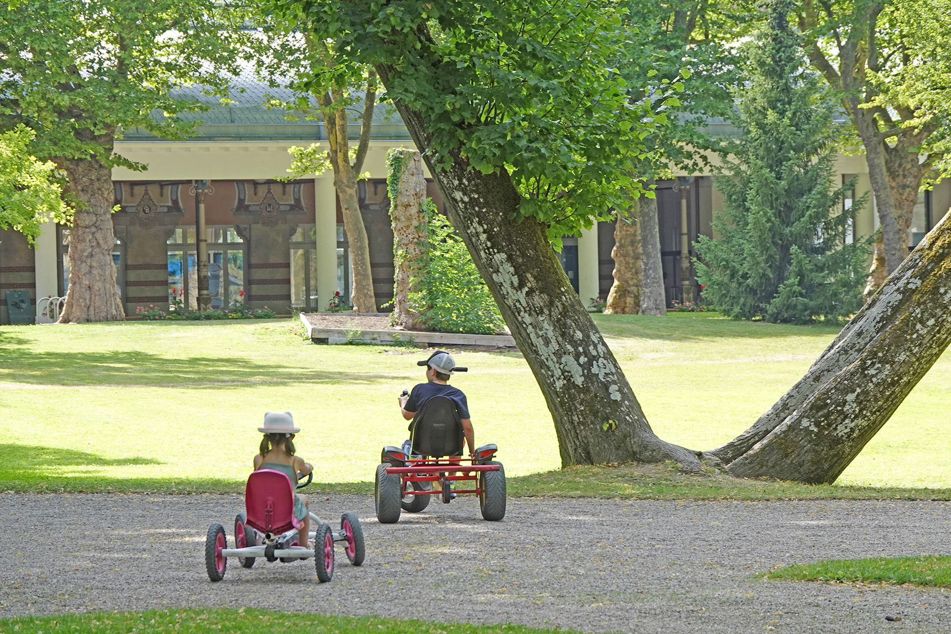 Children ride go-karts on a gravel path in a leafy park with trees and a building in the background at Seasonova Vittel