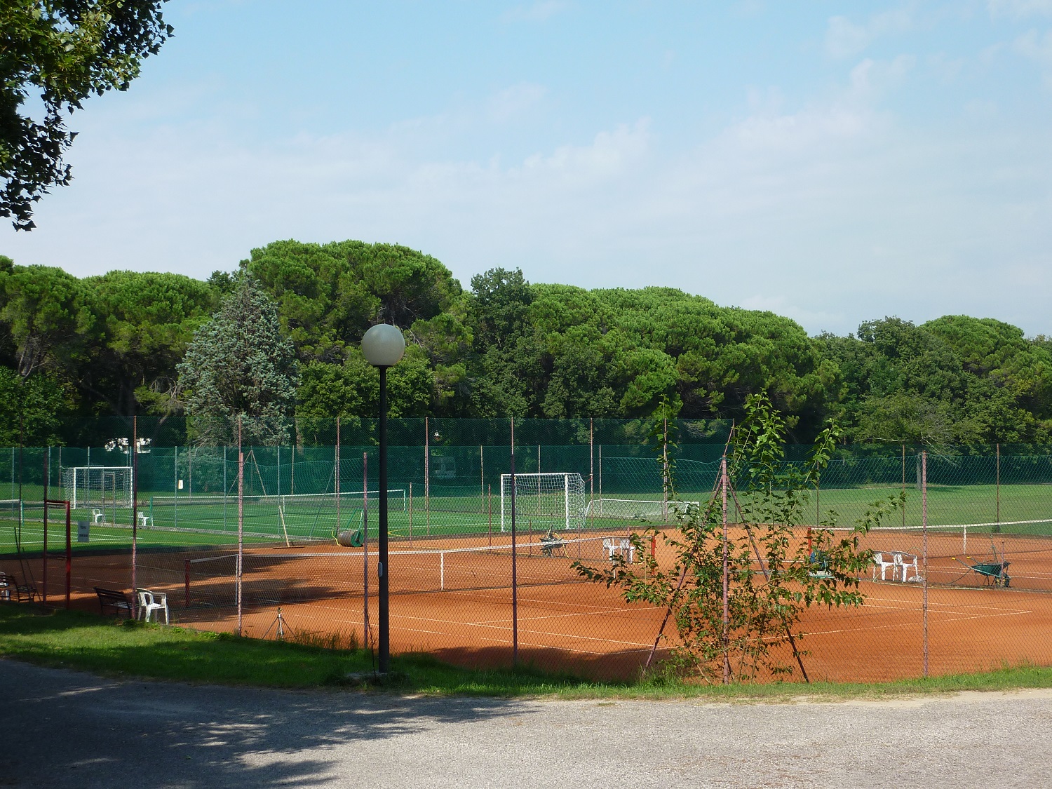 Brown tennis court with chairs and equipment, surrounded by a green fence; lush trees and additional sports fields in the background at Belvedere Pineta
