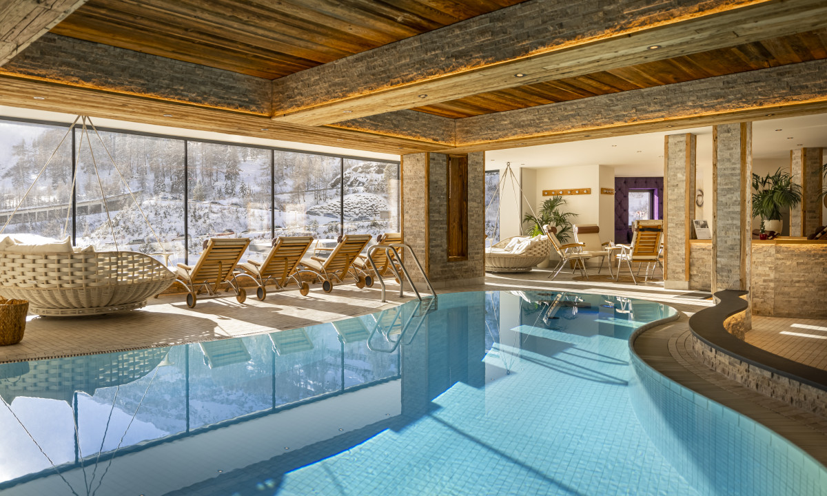 Swimming pool reflects natural light from large windows, surrounded by wooden lounge chairs, overlooking a snowy landscape at Kesselspitze Valamar Collection Hotel