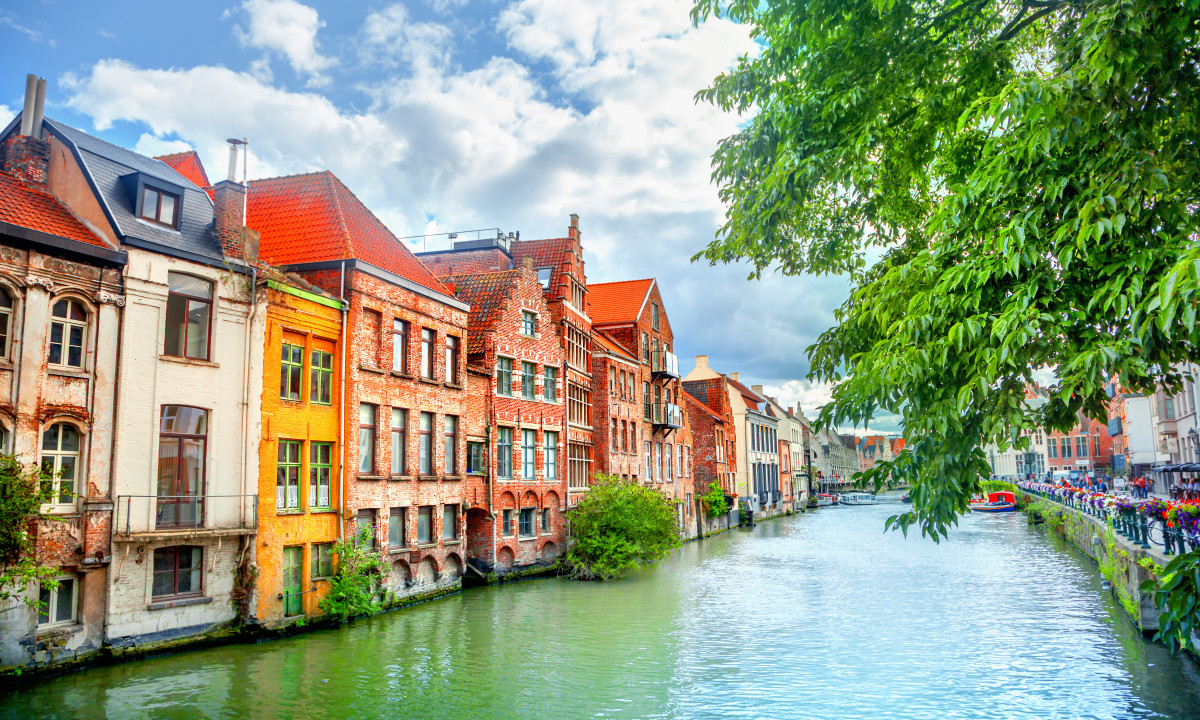 At Urban Gardens Gent, vibrant structures line a serene waterway, bordered by a tree, beneath a partly cloudy sky.