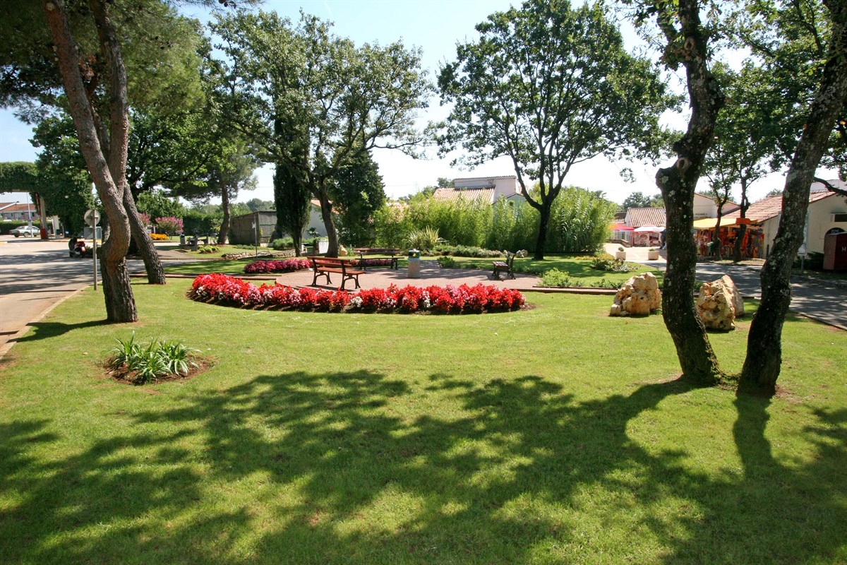 At Naturist Ulika, a verdant park features picnic tables, vibrant flower gardens, and shady trees set against a tranquil residential setting.