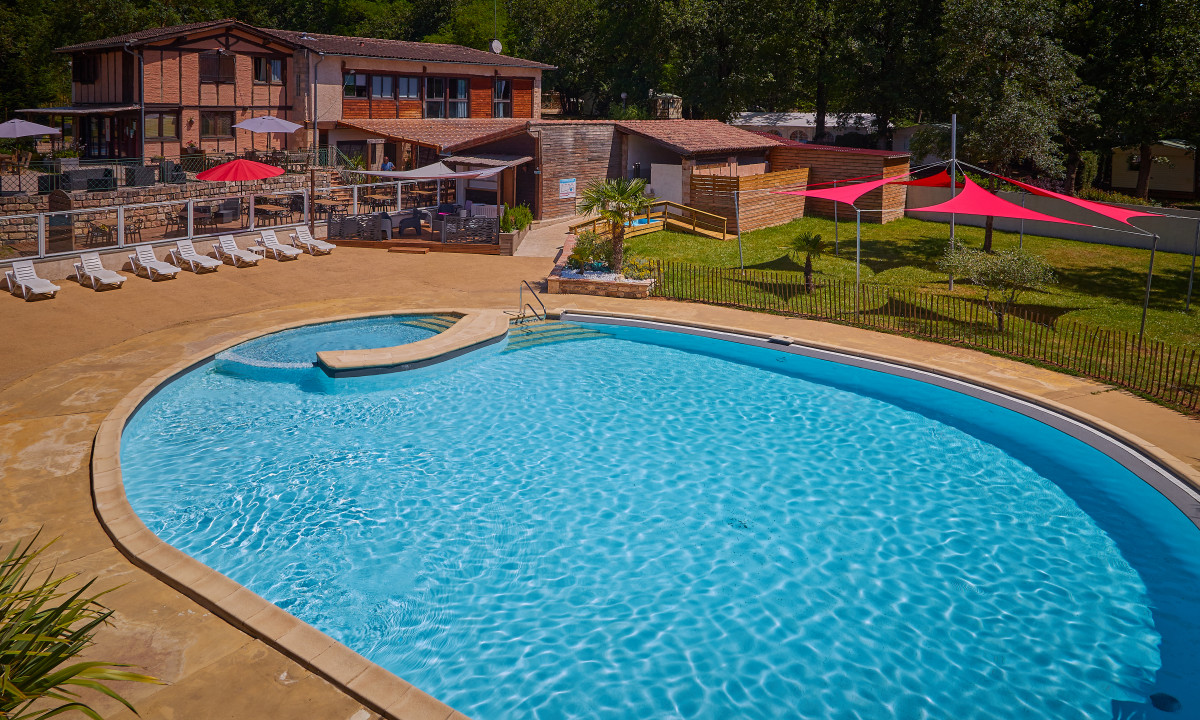 At Le Chêne Vert, there is a spacious swimming pool with crystal-clear water, bordered by sun loungers and shaded spots, adjacent to a structure and landscaped greenery.