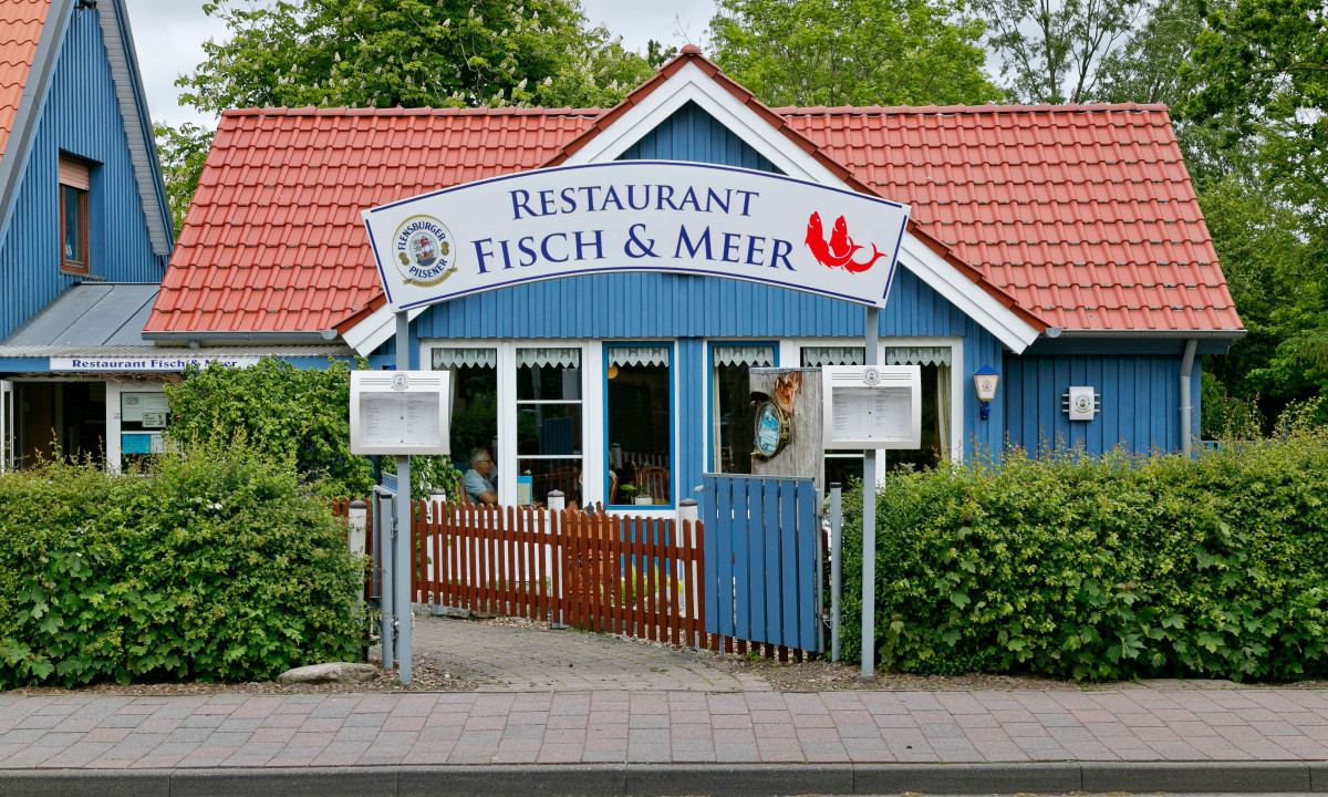 At KNAUS Campingpark Dorum, a blue eatery with a crimson roof showcases menus, encircled by lush greenery on a bright day.