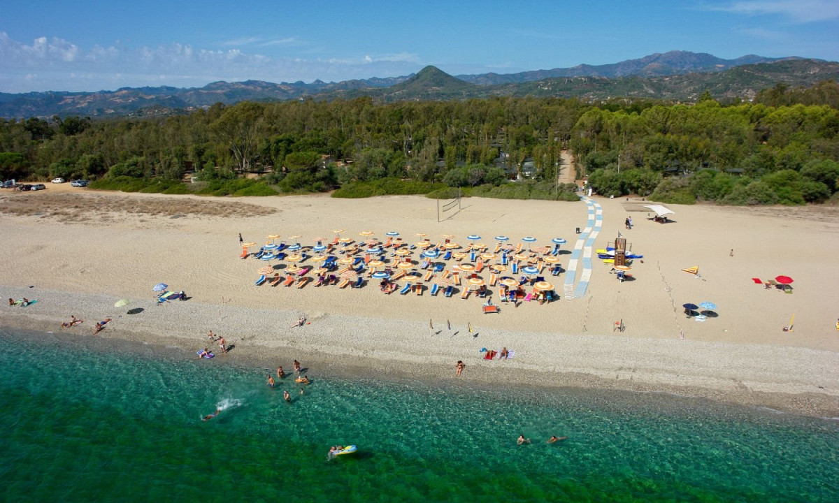 At Ultima Spiaggia, vibrant beach umbrellas and loungers are accompanied by individuals swimming and unwinding on a sandy shore adjacent to lush forests and mountains.