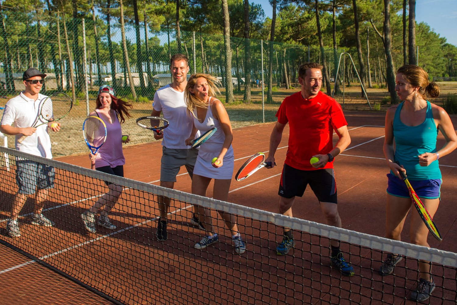 At Marvilla Parks Atlantic Club Montalivet, a group of six individuals is engaged in a game of tennis, positioned on a court with rackets and balls, encircled by trees.