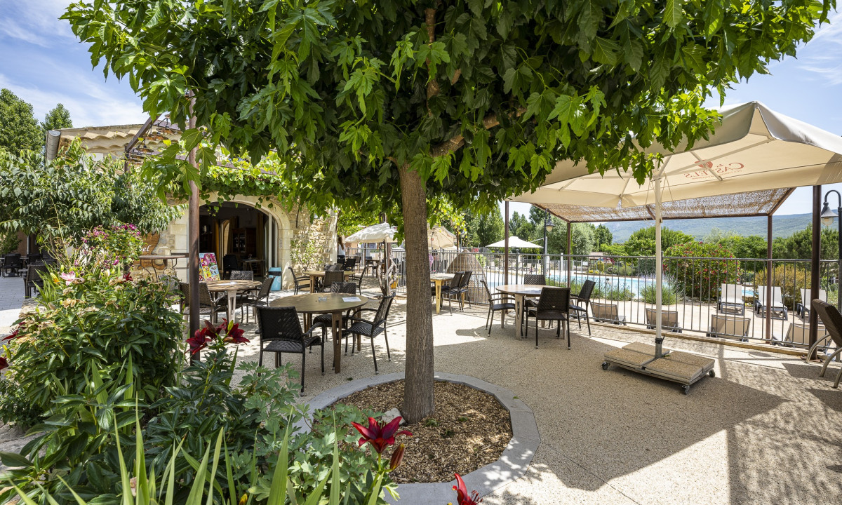 Tables and chairs arranged under a large tree and umbrella near a poolside with scenic mountain views at Domaine de Sévenier