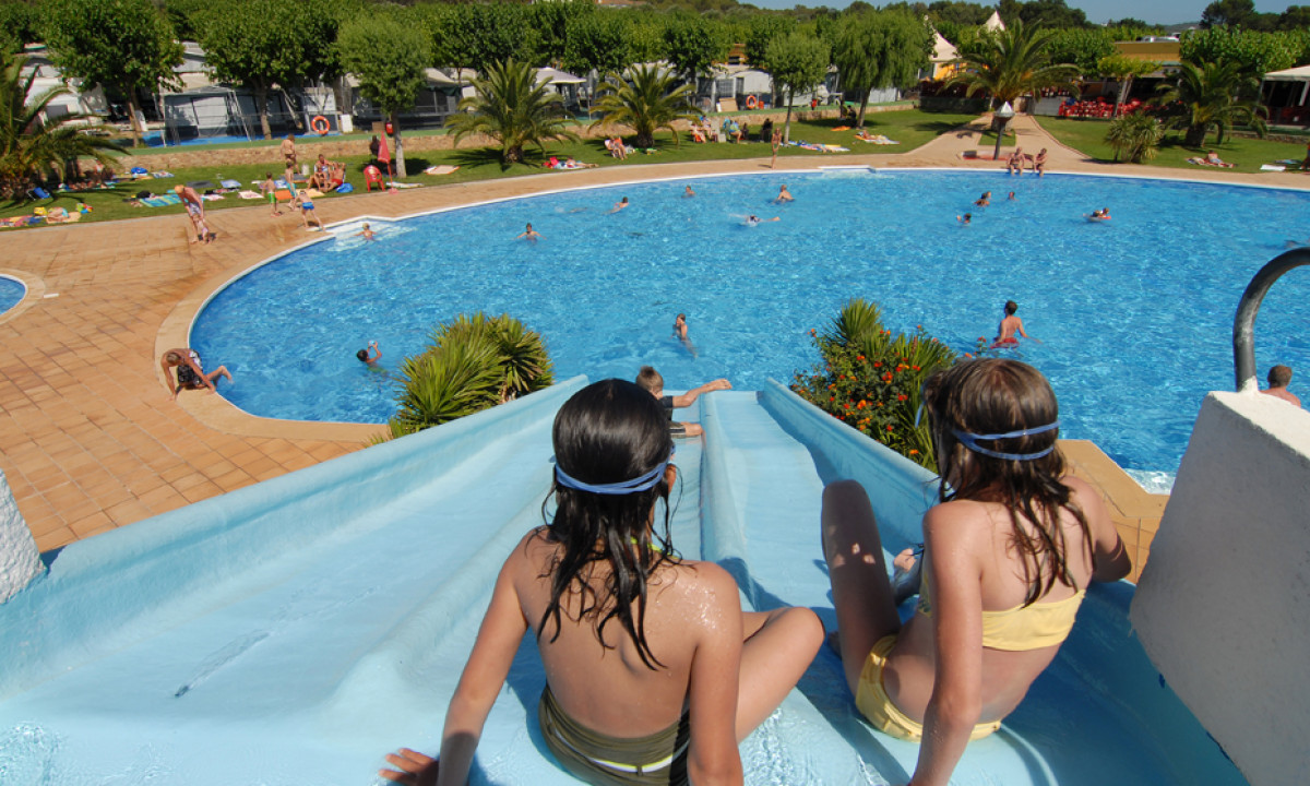 Two girls descend a blue waterslide into a crowded swimming pool surrounded by palm trees and sunbathers at Mas Patoxas