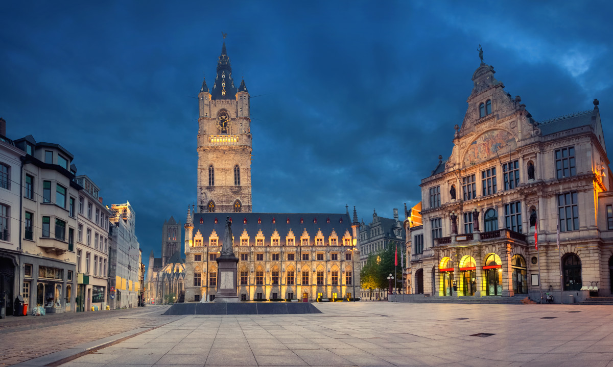 At Urban Gardens Gent, a historic clock tower shines brightly with lights, rising majestically in a town square, encircled by decorative structures.
