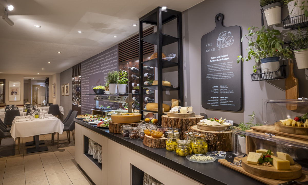 Cheese and fruit display on rustic wooden boards and jars, surrounded by dining tables in a cozy restaurant setting at Valamar Obertauern Hotel