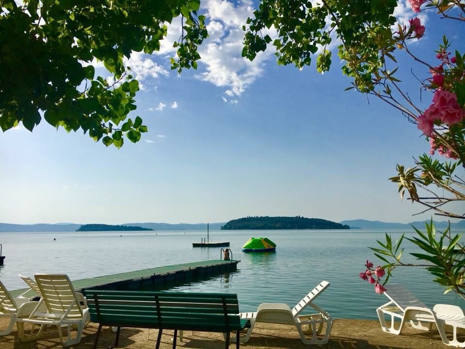 Lounge chairs overlook a calm lake with a wooden dock, inflatable platform, and distant islands, surrounded by foliage at Punta Navaccia