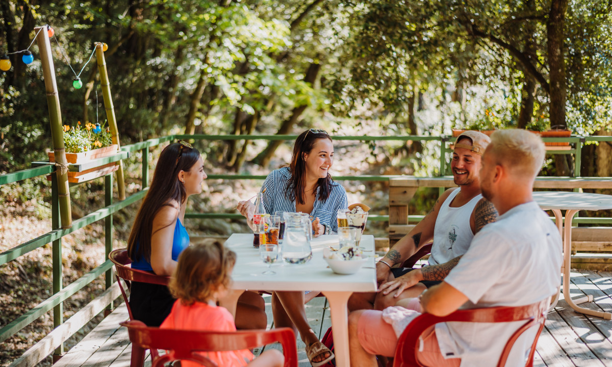 At La Clémentine, individuals gather around a table, chuckling and dining on a wooden patio enveloped by trees.