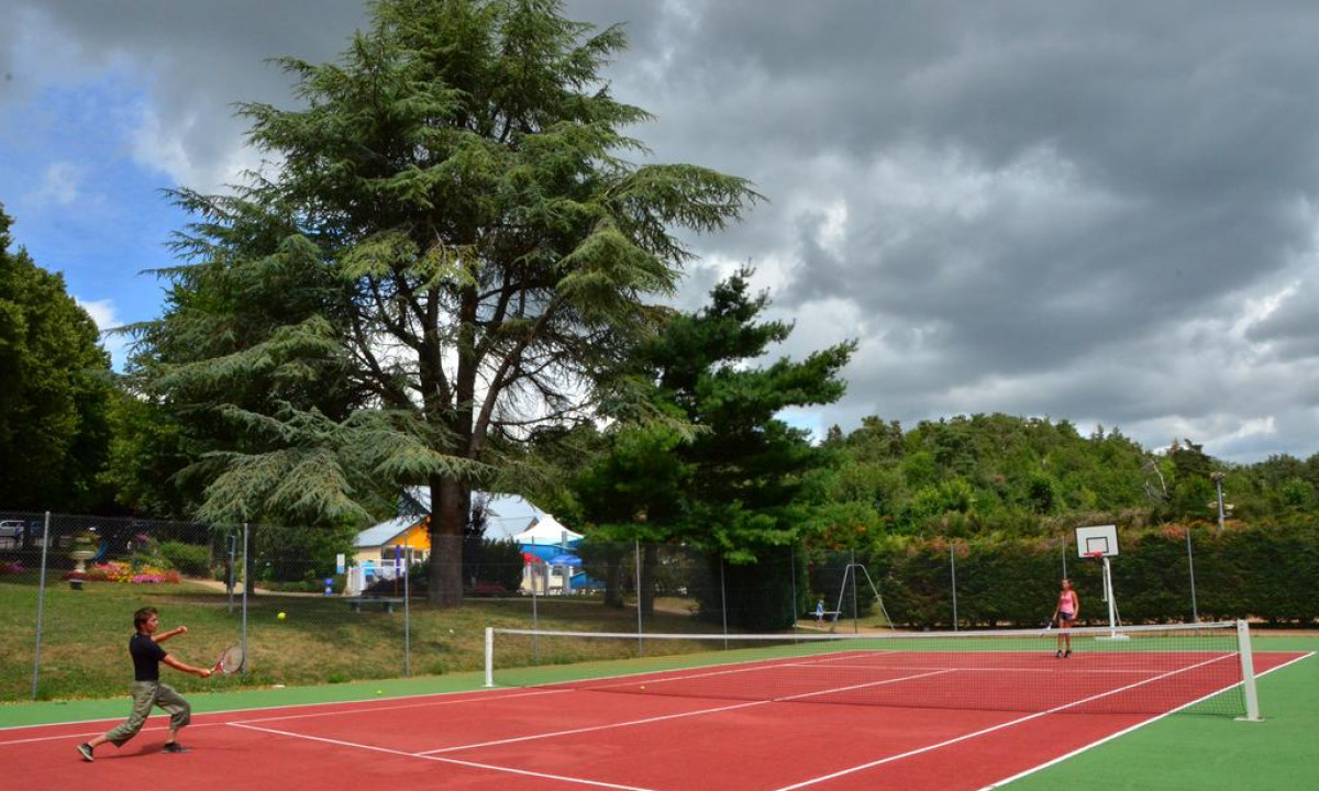 Two people playing tennis on an outdoor red court, surrounded by tall trees and camping facilities at Paradis l'Europe