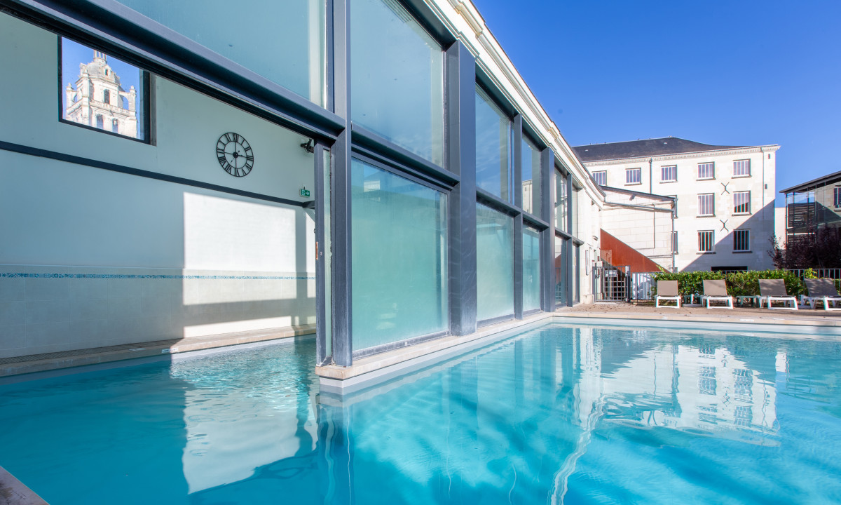Pool spanning indoor and outdoor sections, shaded by building with windows and surrounded by lounge chairs at Le Moulin des Cordeliers