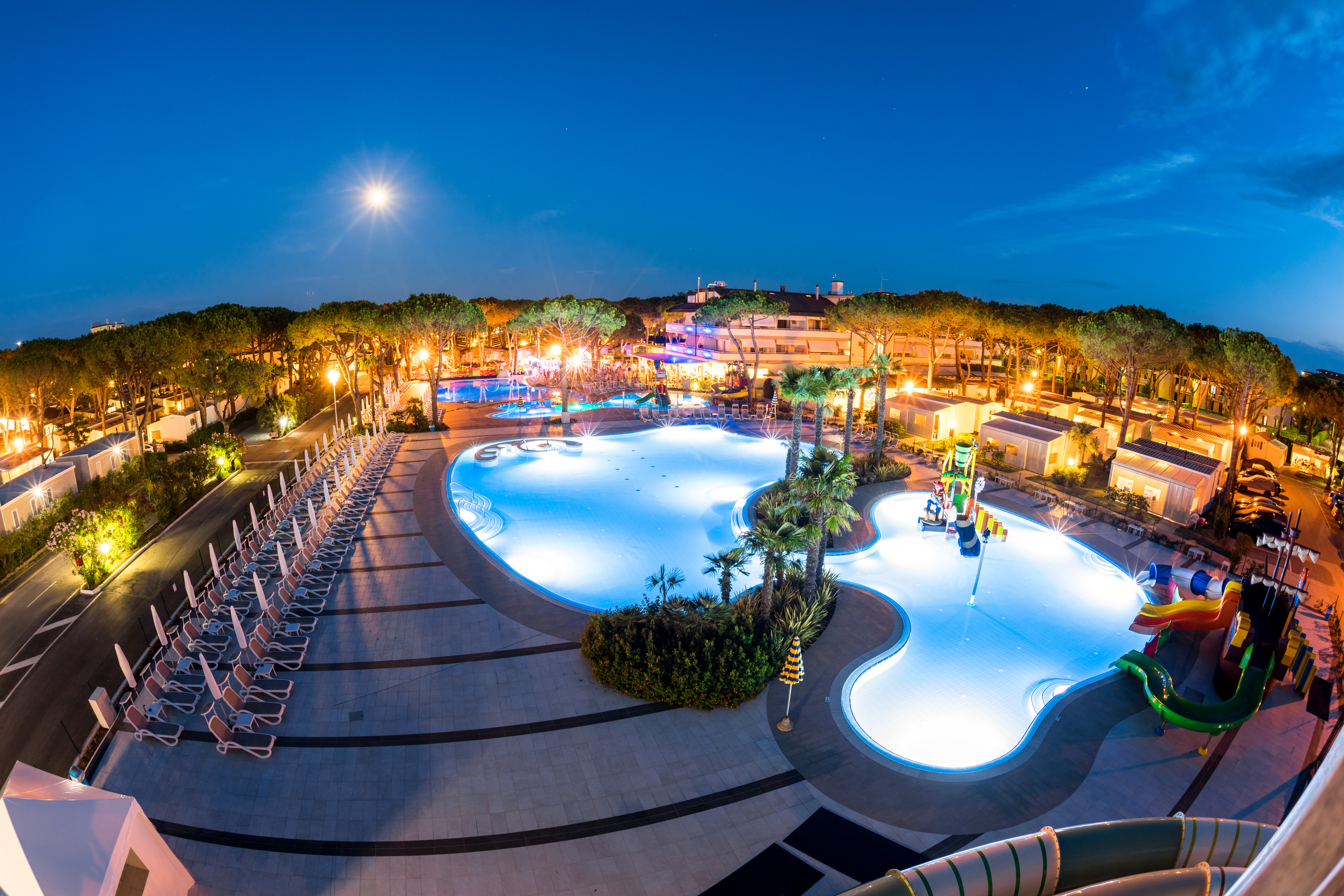 Swimming pool with water slides and deck chairs, surrounded by trees and buildings during evening twilight at Residence Village