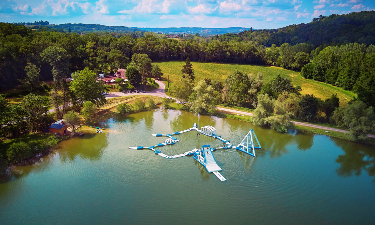 At Coucou, a buoyant obstacle course rests on a lake, encircled by trees and fields beneath a partially cloudy sky.