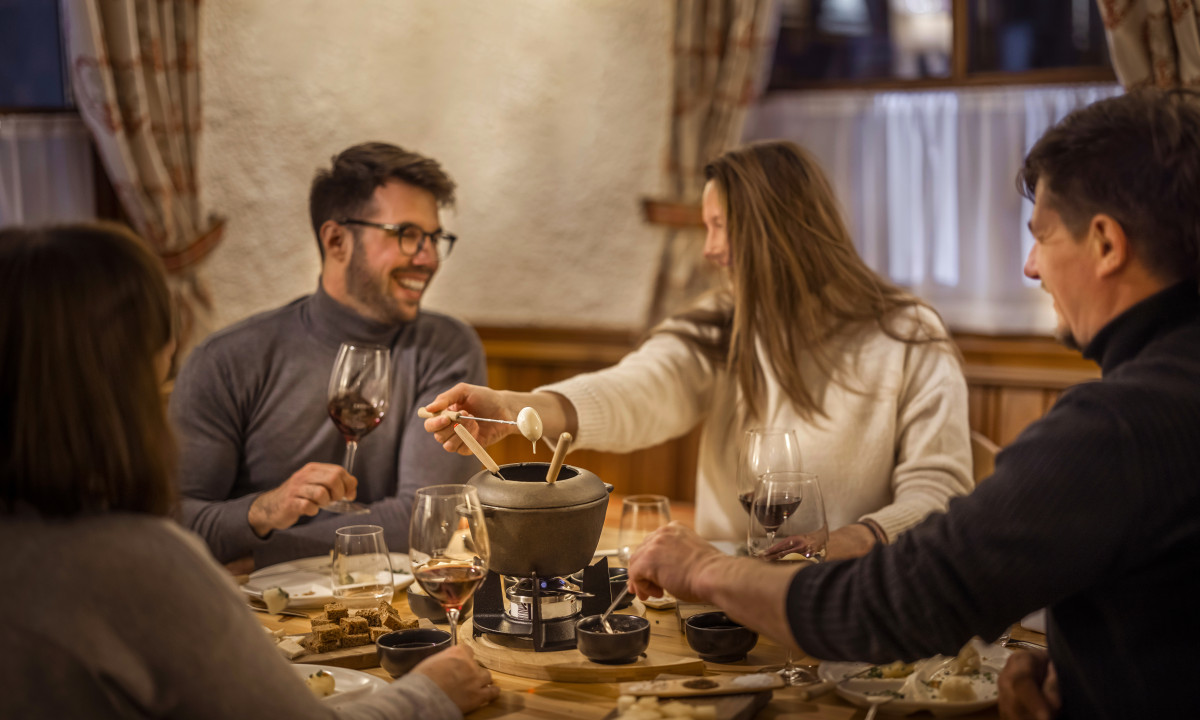 Friends enjoying cheese fondue, dipping food into pot, sitting at a warmly lit dinner table at Valamar Obertauern Hotel
