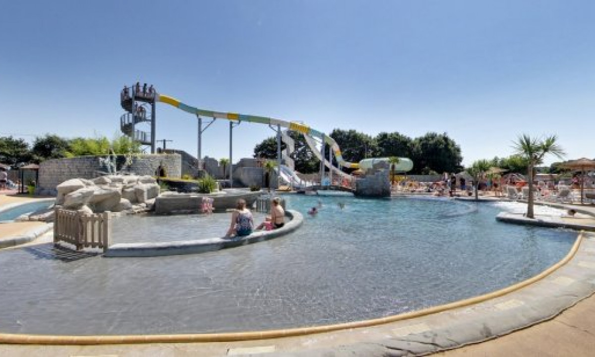 At Le Domaine de Léveno, swimmers enjoy the pool near waterslides while individuals wait on a spiral staircase beneath a clear sky.