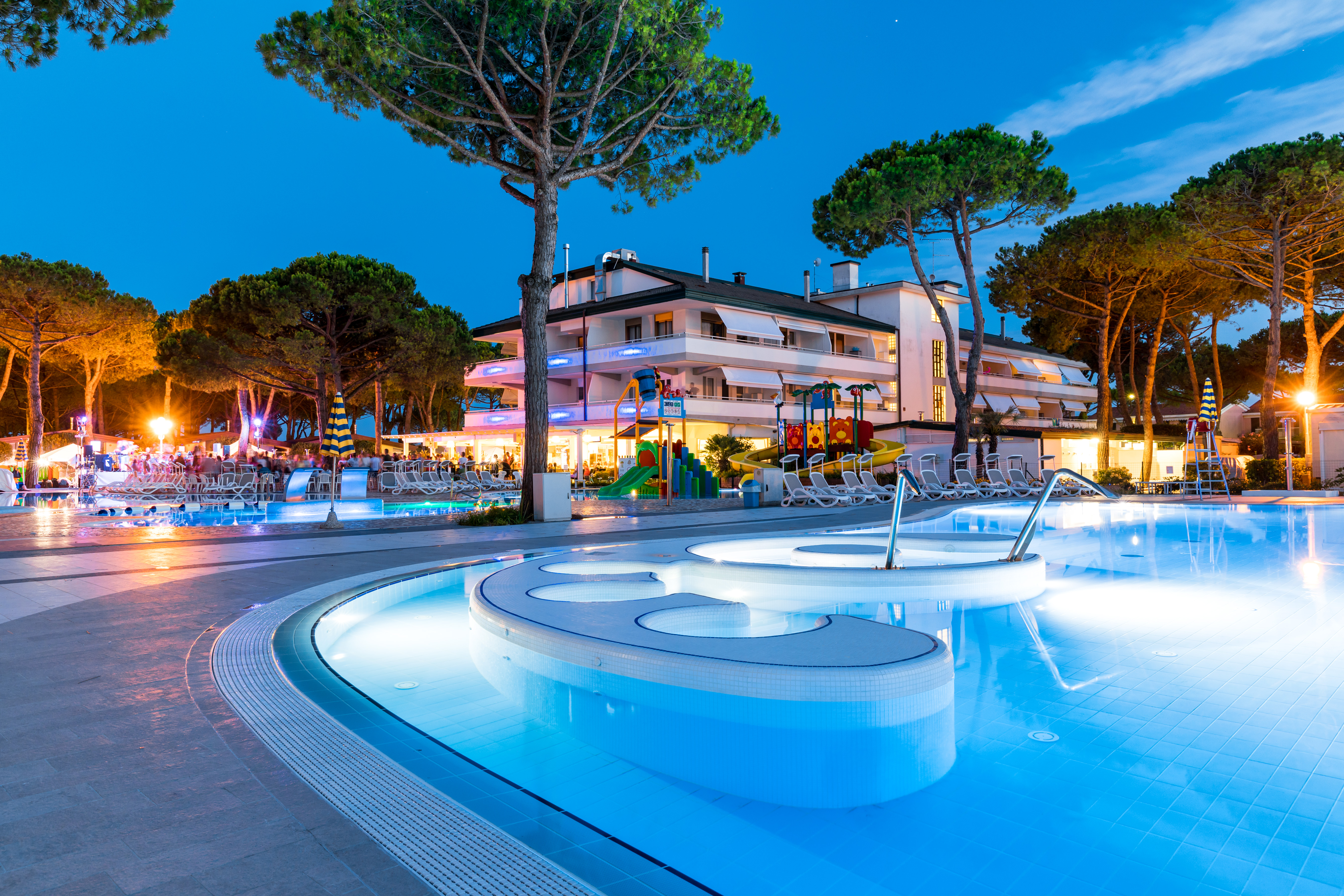 Brightly lit swimming pool beside a lively building with colorful playground and surrounding trees at Residence Village
