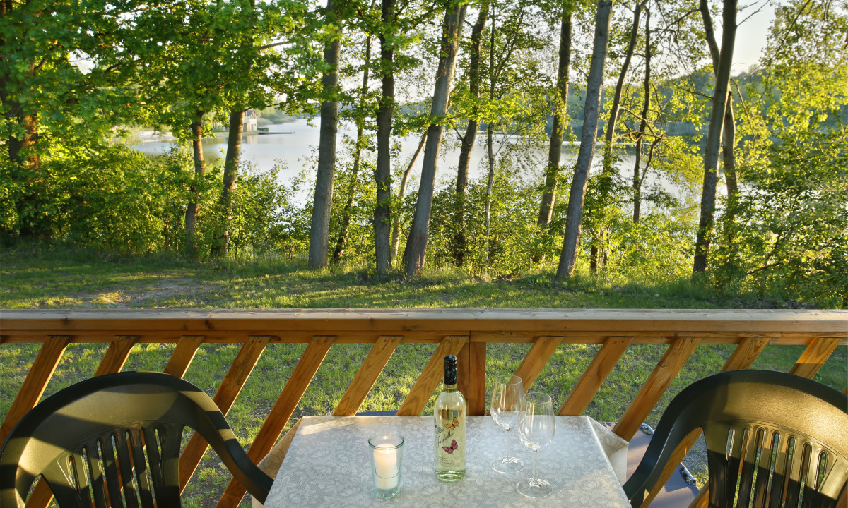 At KNAUS Campingpark Oyten, a table featuring a wine bottle and glasses provides a view of a tranquil lake through the trees.