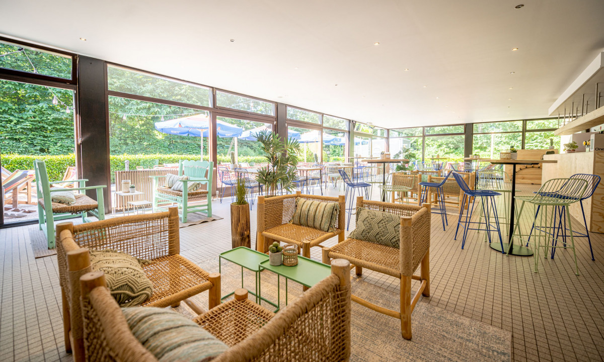 At Urban Gardens Gent, wicker seating and tables are arranged in a vibrant indoor lounge space featuring expansive windows that provide a view of an outdoor patio adorned with umbrellas.