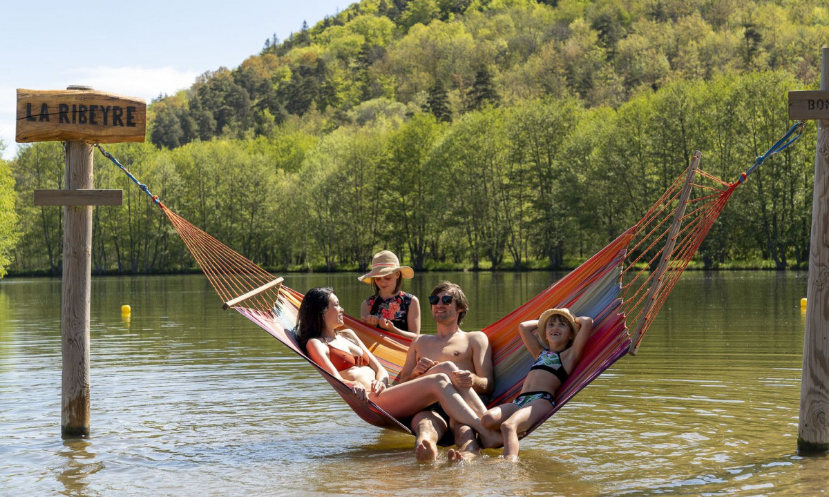 A family relaxes in a hammock suspended over a shallow lake, surrounded by a lush forested hillside at La Ribeyre
