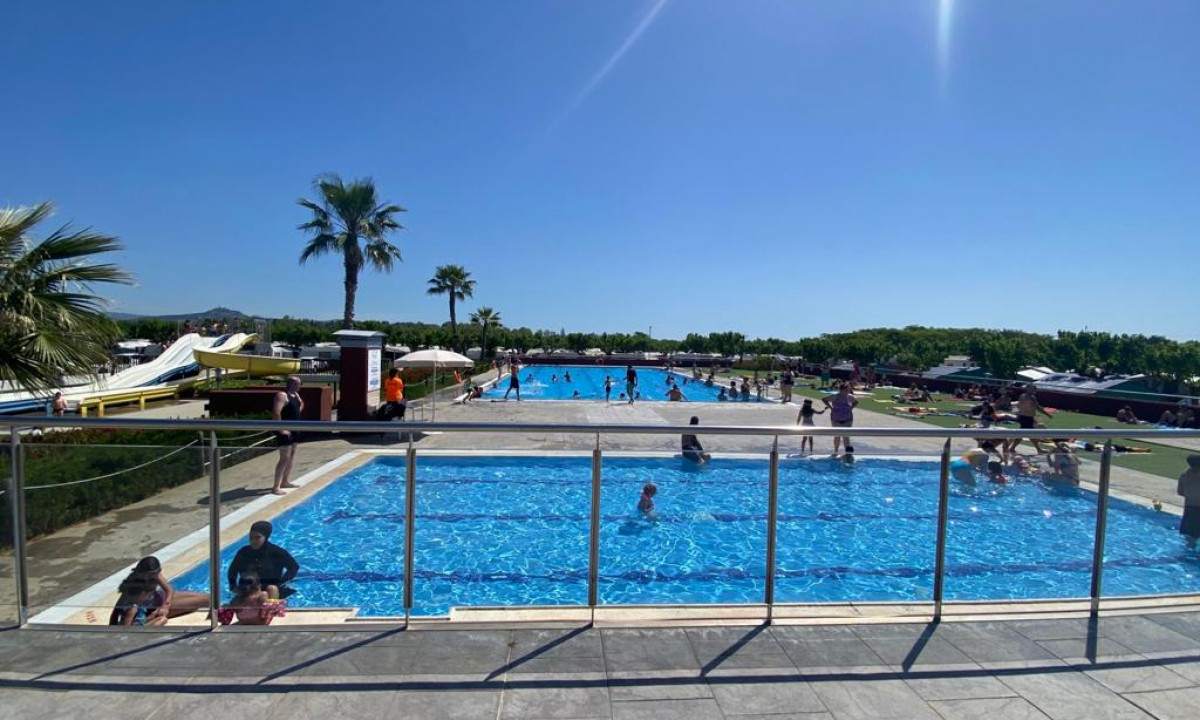 Swimmers enjoying two outdoor pools under a clear sky, with waterslides, palm trees, and sunbathers in the background at Resort Els Pins