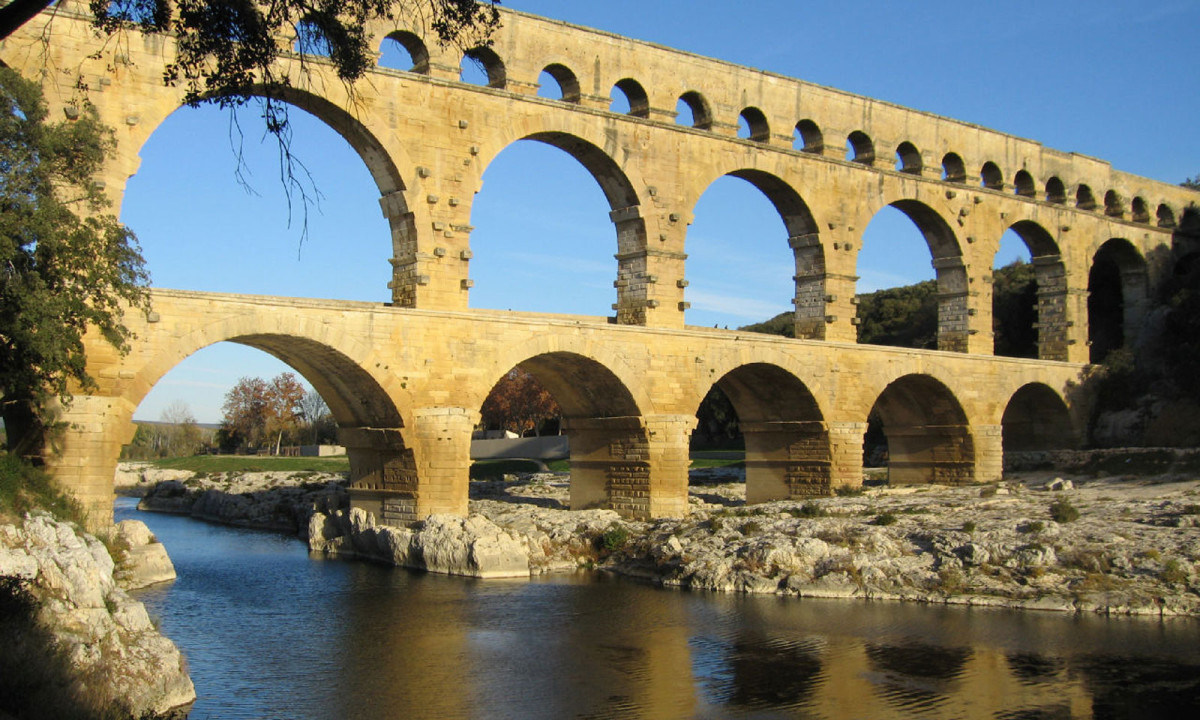 At La Clémentine, ancient stone aqueduct arches span a serene river in a rugged, wooded setting.
