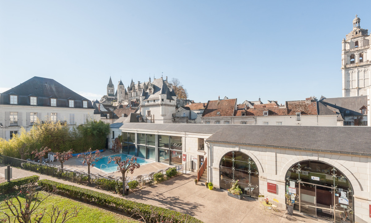 Swimming pool surrounded by buildings with castle and church tower in the background, under a clear blue sky at Le Moulin des Cordeliers