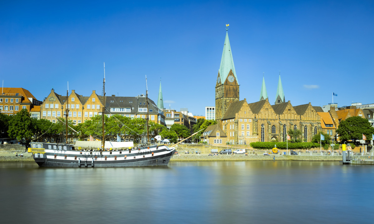 At KNAUS Campingpark Oyten, a sailing vessel is moored in a tranquil river, flanked by historic structures and a church featuring tall spires.