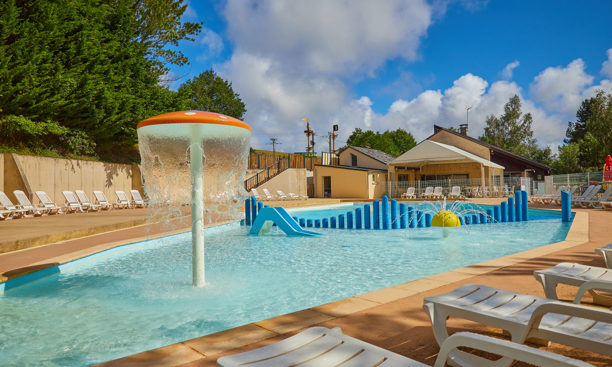 At Terrasses du Lac, there is a children's swimming pool featuring a water fountain and a slide on a sunny day, accompanied by lounge chairs and nearby buildings.