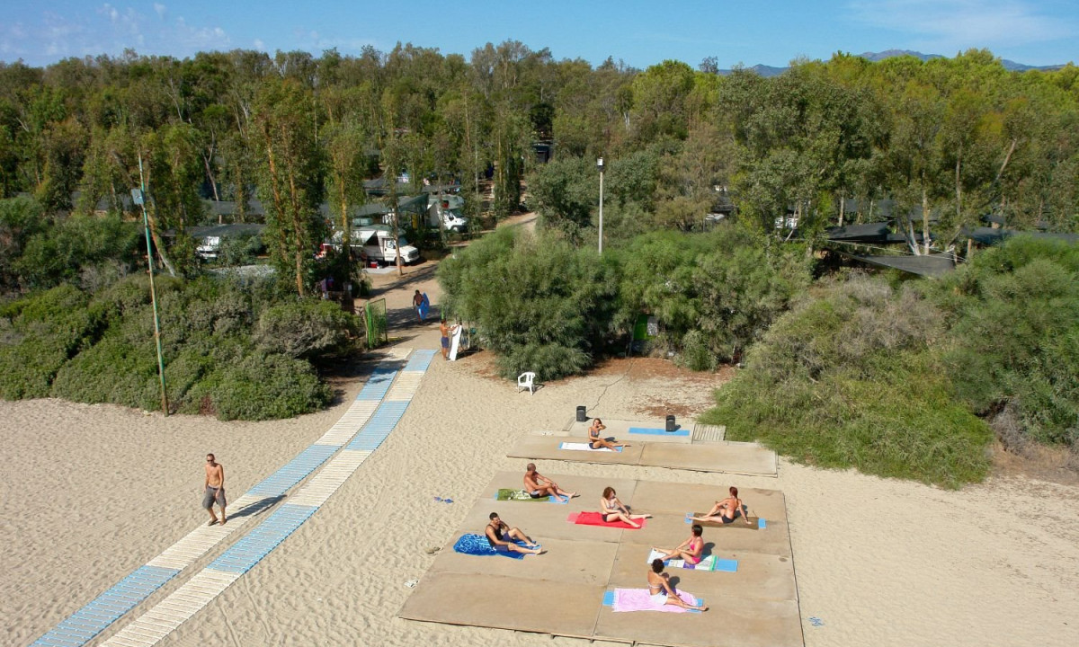 At Ultima Spiaggia, individuals are lounging on a sandy shore, encircled by dispersed towel spots, with trees and parked camper vans in the backdrop.