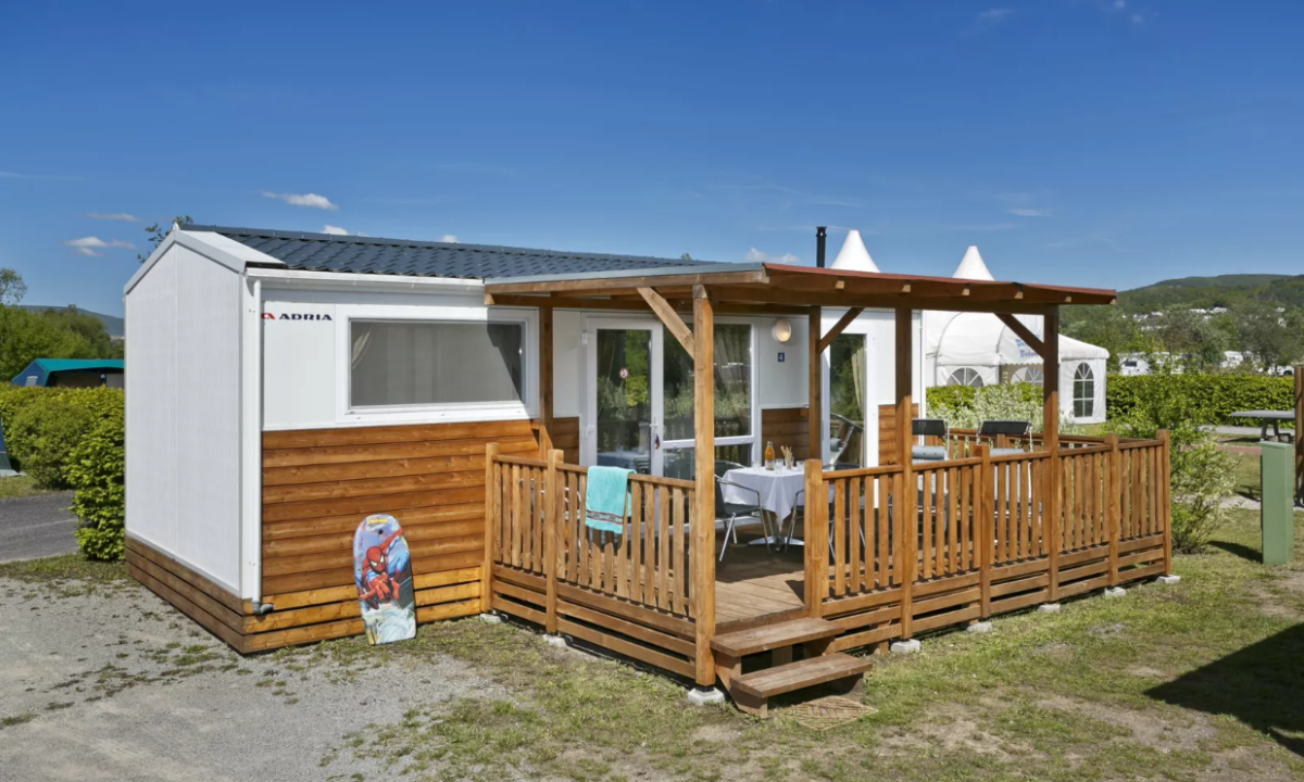 At KNAUS Campingpark Lübben, there is a wooden lodge featuring a cozy porch, a sunbed, and seating, enveloped by lush vegetation and tents.