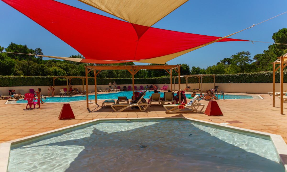 At La Sousta, individuals unwind by the pool on lounge chairs beneath vibrant shade sails, encircled by trees and a clear blue sky.