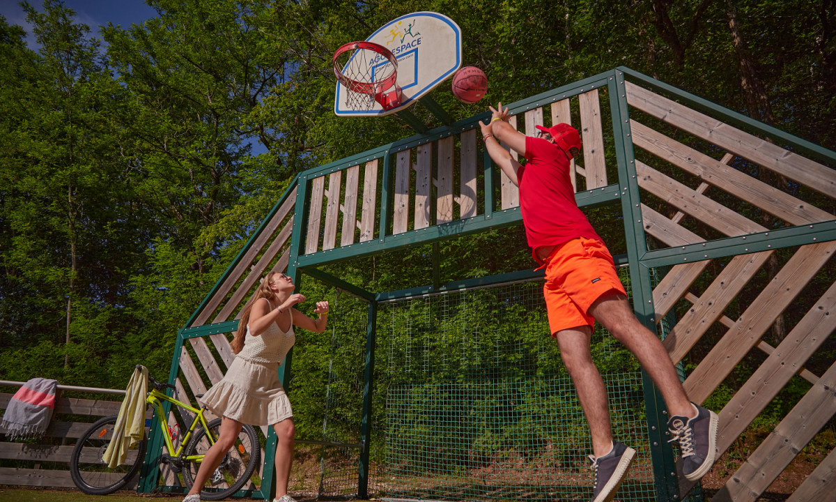 At the outdoor court surrounded by trees, two individuals are playing basketball, with one player leaping to take a shot at Coucou.