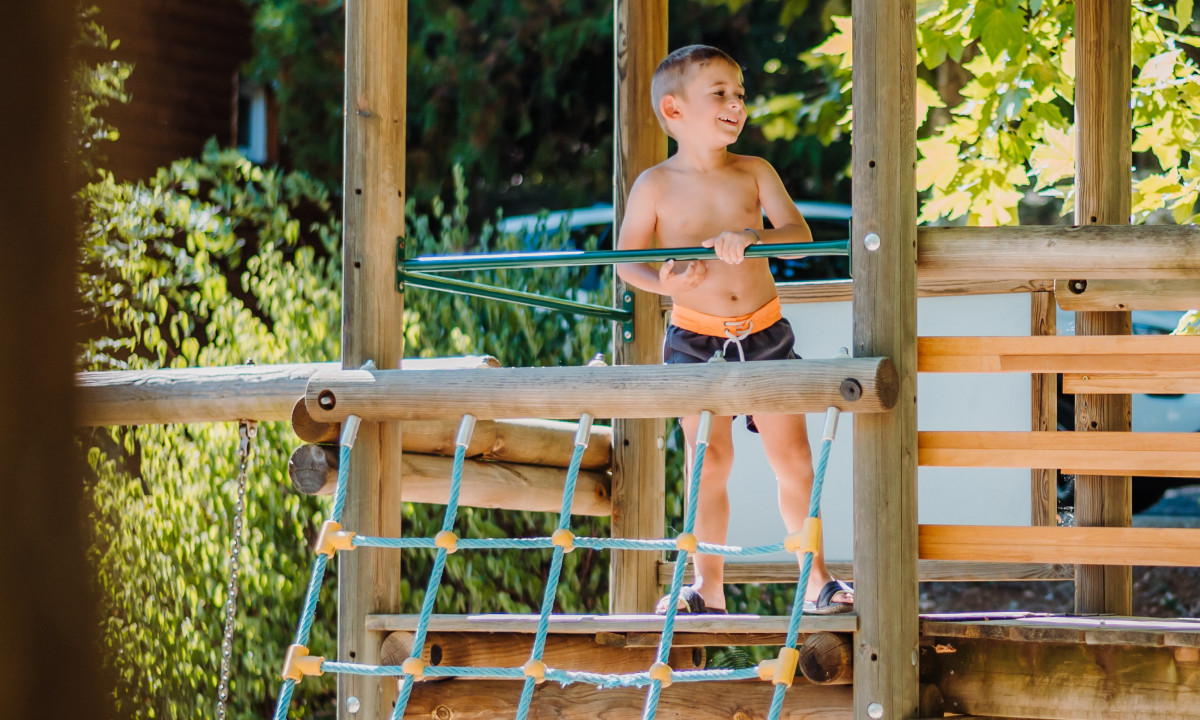 At La Clémentine, a child is positioned on a wooden play structure, grinning, encircled by lush greenery and trees.