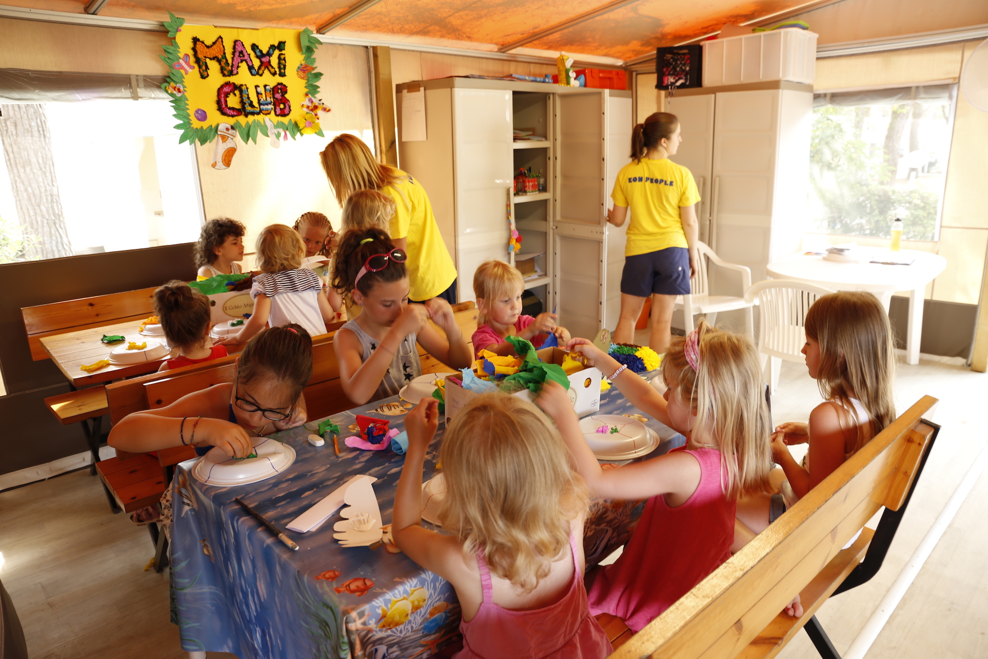 Children crafting at a table while staff assist in an indoor campsite play area for kids at Residence Village